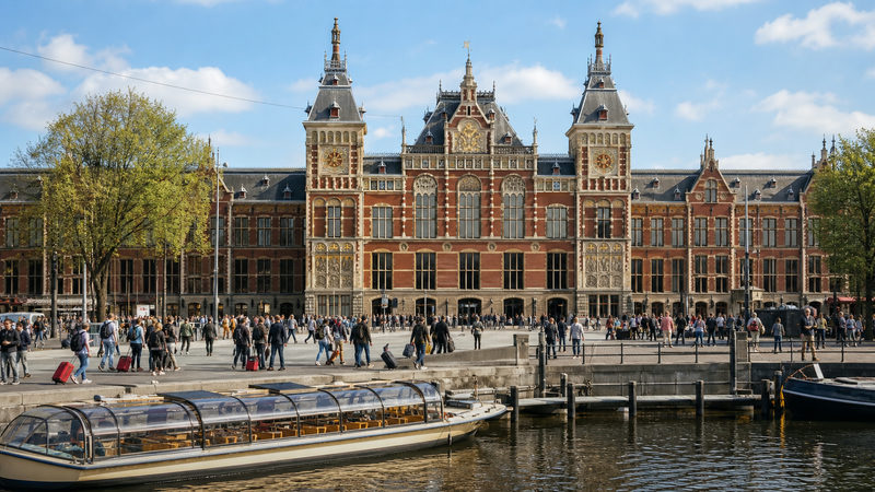 Voorgevel van Amsterdam Centraal Station gezien vanaf het Stationsplein, met reizigers met koffers op de voorgrond en het IJ op de achtergrond
